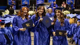 Three women dressed in blue graduation cap and gowns hold up TCC degrees