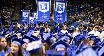 Two students in blue graduation regalia each hold a TCC gonfalon in a crowd of graduating students