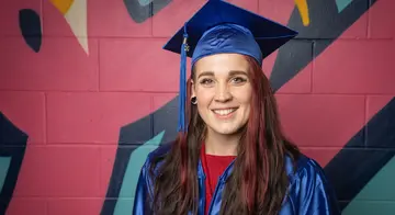 Lizz West wears blue graduation regalia and stands in front of a brightly colored wall
