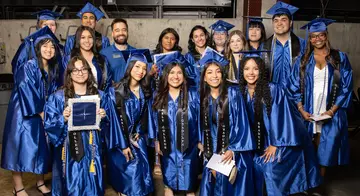 A group of 16 concurrent graduates smile in their regalia before TCC's Spring 2026 Commencement Ceremony. A TCC Dual Credit employee smiles with them.