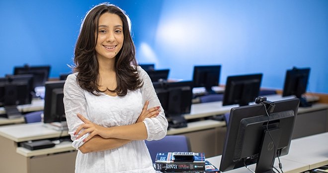 Young woman smiles with arms crossed, standing in a computer lab at TCC.