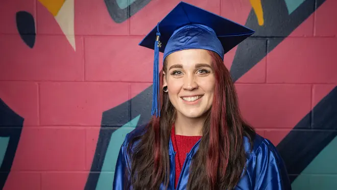 Lizz West wears blue graduation regalia and stands in front of a brightly colored wall