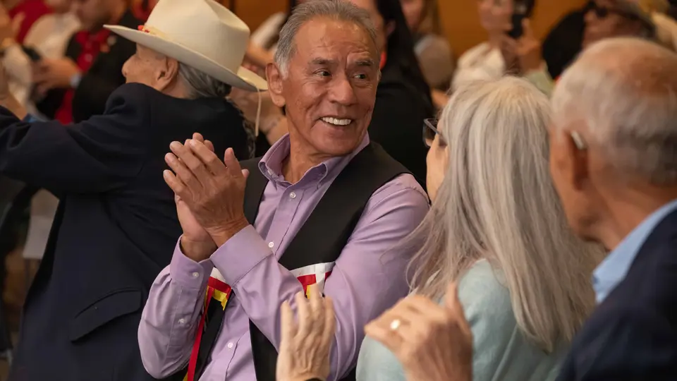 Wes Studi is sitting down and smiling in a crowd of people.