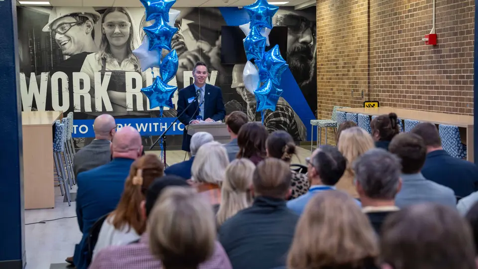 Dr. Kristopher Copeland stands behind a podium flanked by blue and white balloons in the entrance of the Engineering Technology Building. He addresses a crowd of sitting people.