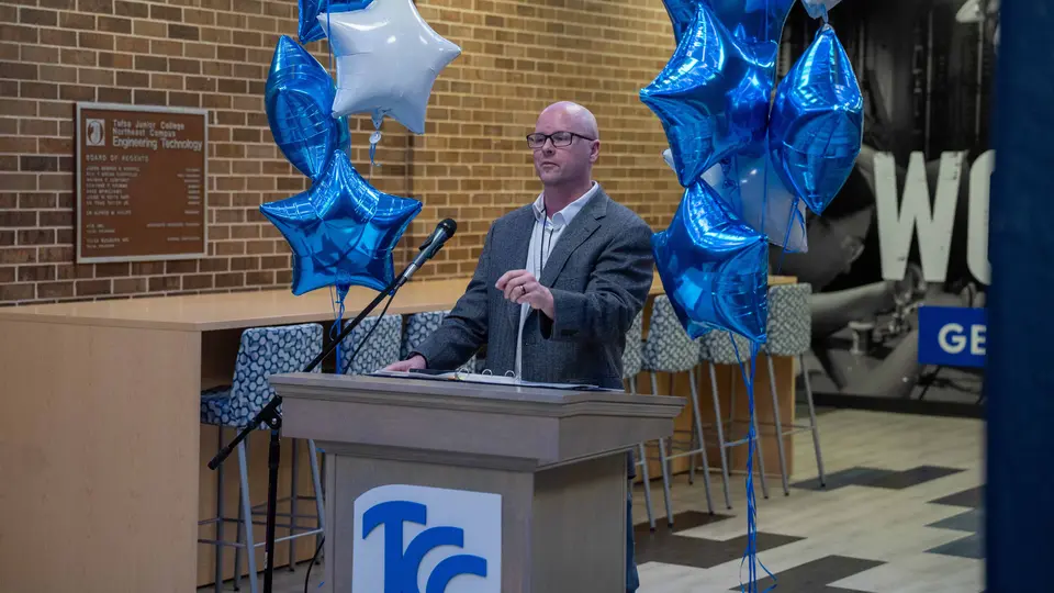 Dr. David Paige speaks behind a podium flanked by blue and white balloons inside the Engineering Technology Building.
