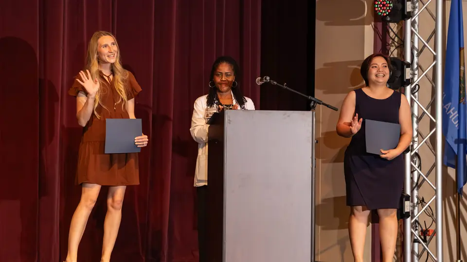 A faculty member stands at a podium on the VanTrease PACE stage with two female students on either side of her.