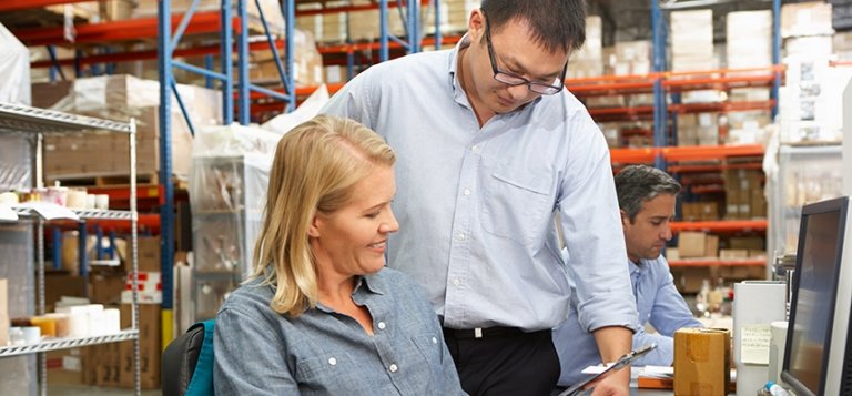 Asian man stands next to seated white woman. Both review information on a tablet.