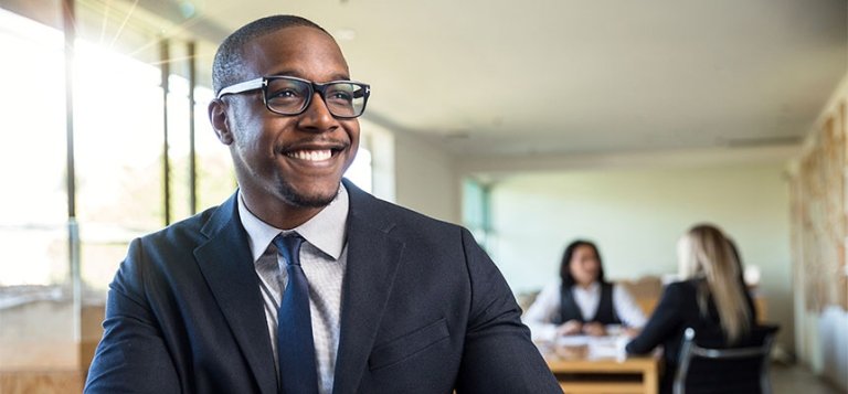 Young black man smiles in a bright conference room with people sitting at the table in the blurred background.