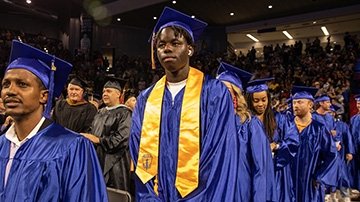 Black male TCC graduate wears blue graduation cap and gown with gold Phi Theta Kappa Honors Society sash.