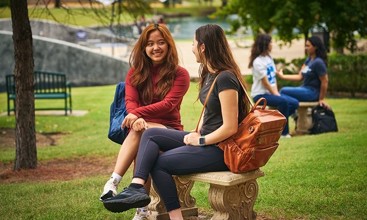 Two TCC Students sit on a bench outside under trees at the Southeast campus.