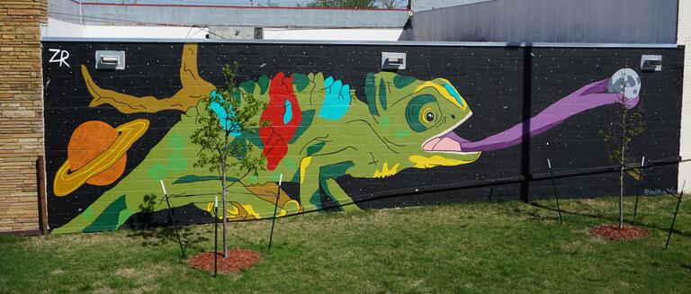 'Caught in Space' shows a large green chameleon in space. Its tongue is outstretched and grabbing a planet or space rock
