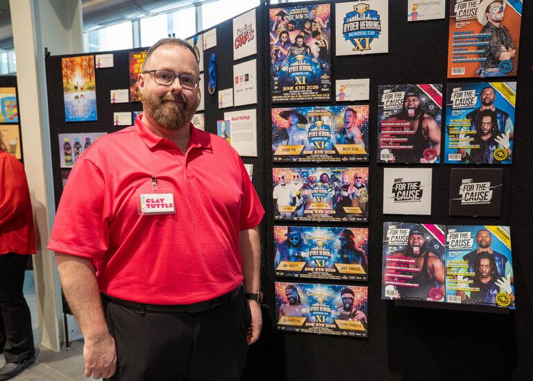 Student Clay Tuttle stands next to his display, which includes several magazine covers for the nonprofit organization Wrestling for a Cause. The magazine covers feature photos of different wrestlers.