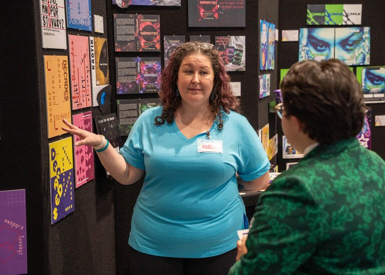 Digital Media student MaryAnn Ferrell points to a poster design on her display while she talks to another person.