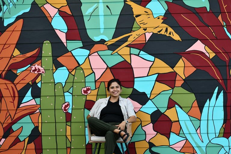 Zadith sits in front of a bright mural that is at the Brut Hotel. The mural shows a green cactus, yellow scissortail flycatcher, and various brightly colored plants and geometric shapes.