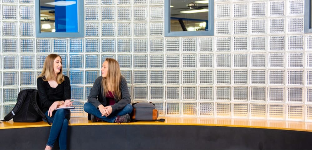Two Tulsa Community College students chatting on a bench in front of a wall of glass bricks